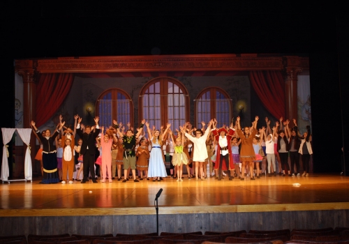 The full FPAS cast on stage with arms raised at the end of a summer camp performance